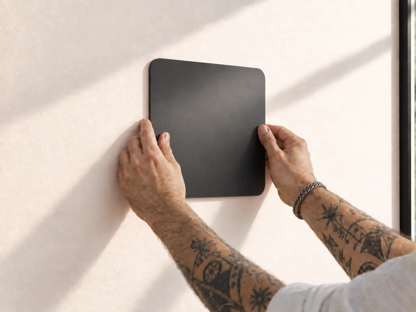 Two hands pressing a black square mount flat against a pale plaster wall in low sunlight